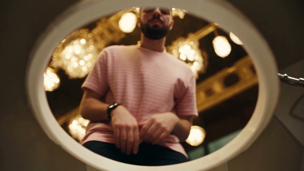 Source frame of a man in a striped shirt seen through an oval mirror, gilded chandeliers reflected behind him