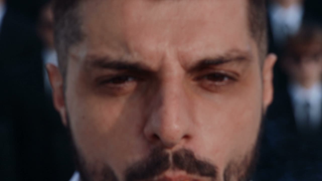 Close-up source frame of a man's furrowed brow, formally dressed figures blurred in the background